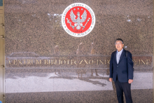Mężczyzna stoi przy ścianie z godłem Warszawskiego Uniwersytetu Medycznego i napisem „Centrum Biblioteczno-Informacyjne”. Zdjęcie wykonano przed wejściem do budynku.