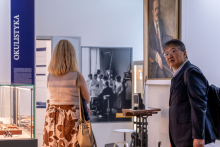 A woman and a man are in an exhibition space dedicated to the history of medicine. One person is viewing the exhibition, while the other is walking by. In the background, display cases, informational panels, and historical photographs are visible.