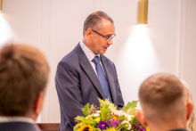 Mężczyzna przemawia do uczestników spotkania. Na pierwszym planie widoczne są osoby siedzące przy stołach oraz kompozycja kwiatowa.