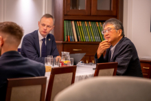Kilka osób uczestniczy w spotkaniu przy stole konferencyjnym. W tle znajduje się regał z publikacjami, a obok widoczny jest ekran prezentacyjny.