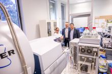 Trzy osoby oglądają aparaturę badawczą w laboratorium. W kadrze widoczne są urządzenia analityczne, komputery oraz wyposażenie pracowni.