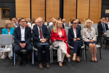Uczestnicy wydarzenia siedzą w pierwszym rzędzie sali konferencyjnej. W tle widoczni są pozostali goście zgromadzeni na spotkaniu.