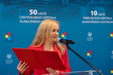 Osoba występuje przy mównicy, trzymając otwartą teczkę z dokumentami. W tle widoczne są materiały promocyjne UCK WUM.