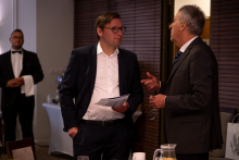 Dwóch mężczyzn w garniturach rozmawia podczas spotkania przy stole konferencyjnym. Jeden z nich trzyma w ręku dokumenty. W tle stoi kelner ubrany w smoking i muszkę.
