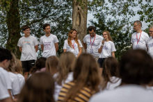 Młoda kobieta trzyma mikrofon i przemawia do uczestników spotkania. Obok stoją kobiety i mężczyźni należący do zespołu organizacyjnego.