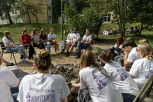 Uczestniczki i uczestnicy siedzą w kręgu wokół miejsca na ognisko i biorą udział w dyskusji prowadzonej przez organizatorów.