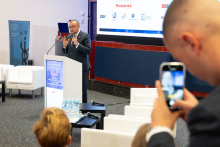 Conference presentation observed by attendees while one participant records the speech using a smartphone.