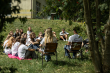 Grupa studentek i studentów siedzi na trawie w cieniu drzew podczas spotkania warsztatowego na świeżym powietrzu.