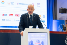 A speaker addresses the audience from a podium in front of a screen displaying the conference theme and logos of organizers and partners.