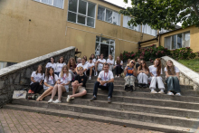 Participants seated on outdoor steps listening to a presentation near a building entrance.