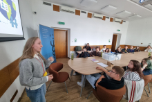 Prowadząca prowadzi zajęcia edukacyjne przy stole konferencyjnym. Kilkanaście osób uczestniczy w spotkaniu i słucha wyjaśnień.