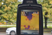 Ekran kamery termowizyjnej prezentuje rozkład temperatury badanego obiektu. W tle widoczne są drzewa i zaparkowany samochód.