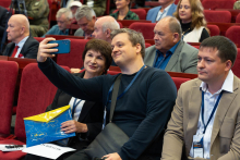 Conference participants seated in an auditorium take a smartphone photo during a conference session.