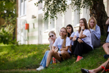 Small group of participants sitting on grass under a tree beside a building, listening during an outdoor event.