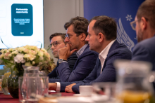 Uczestnicy spotkania siedzą przy stole konferencyjnym i słuchają prezentacji. Na stole znajdują się dzbanki z napojami, filiżanki oraz kompozycja kwiatowa.
