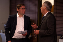 Two participants in business attire converse during a networking session, with one holding printed documents.