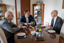 Trzech mężczyzn siedzi przy stole i prowadzą rozmowę podczas spotkania. Na stole stoją napoje oraz talerze z przekąskami.