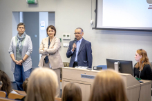 Kobieta stoi pomiędzy rzędami siedzeń w sali wykładowej i przemawia do uczestników, trzymając mikrofon w dłoni.