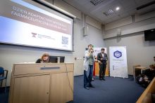 Kobieta, kobieta i mężczyzna stoją na scenie podczas Dnia Adaptacyjnego Wydziału Farmaceutycznego WUM. Kobieta trzyma mikrofon w dłoni, a pozostali prowadzący trzymają kartki z notatkami.