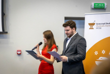 Kobieta i mężczyzna stoją obok siebie podczas prowadzenia wydarzenia. Kobieta trzyma mikrofon i kartkę z notatkami, mężczyzna trzyma kartki w dłoniach.