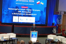 Jedna osoba przemawia przy mównicy podczas konferencji „Samorząd w czasie wojny i kryzysu. Jak skorzystać z doświadczeń ukraińskich?” na Warszawskim Uniwersytecie Medycznym. W pierwszych rzędach siedzą uczestnicy wydarzenia, a za prelegentem widoczny jest ekran z grafiką konferencji.