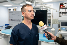A physician gives an interview to the media in a procedure room equipped with modern diagnostic equipment.