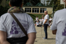 A speaker holding a microphone addresses participants gathered outdoors near a sports court.
