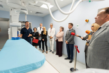 A group of event participants listens to a presentation in a hospital room equipped with medical equipment. Media representatives are present.