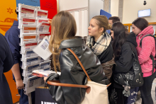 Kilka osób przegląda materiały informacyjne umieszczone na stojaku przy stoisku Warszawskiego Uniwersytetu Medycznego podczas wydarzenia informacyjnego.