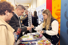 Osoba obsługująca stoisko Warszawskiego Uniwersytetu Medycznego przekazuje materiały promocyjne odwiedzającym. Na blacie znajdują się ulotki, przypinki i smycze.