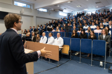 Mężczyzna przemawia do uczestników zgromadzonych w auli. W pierwszych rzędach siedzą studenci oraz przedstawiciele uczelni.