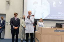 Mężczyzna w białym fartuchu przemawia do uczestników wydarzenia. W tle stoją członkowie zespołu organizującego ceremonię.