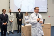 Mężczyzna w białym fartuchu przemawia do uczestników wydarzenia. W tle stoją członkowie zespołu organizującego ceremonię.