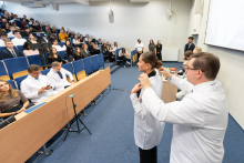 Mężczyzn w białym fartuchu zakłada biały fartuch studentce w czasie ceremonii. W tle widać innych uczestników wydarzenia siedzących w rzędach na auli.