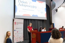 Kobieta przemawia przy mównicy podczas Dnia Adaptacyjnego. W tle widoczny jest ekran z grafiką wydarzenia oraz osoby siedzące przy stole prezydialnym.