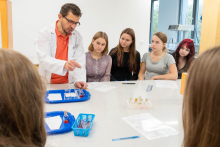 Prowadzący prezentuje doświadczenie chemiczne przy stole laboratoryjnym grupie uczestników obserwujących przebieg ćwiczenia.