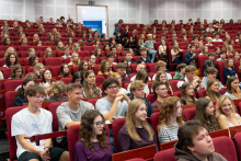 Studentki i studenci siedzą w rzędach czerwonych foteli auli. Część osób trzyma w dłoniach telefony komórkowe i fotografuje lub obserwuje wydarzenie.