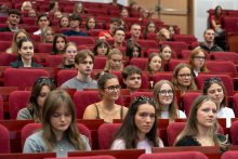 Studentki i studenci siedzą w rzędach czerwonych foteli auli. Kilka osób trzyma w dłoniach telefony komórkowe.