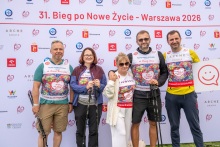 Five participants pose for a photo in front of a backdrop promoting the “31st Run for New Life, Warsaw 2026.” The participants stand in a row and hold Nordic walking poles.