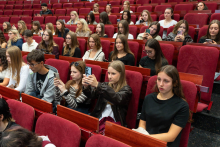 Studentki i studenci siedzą w rzędach czerwonych foteli auli. Kilka osób trzyma w dłoniach telefony komórkowe.