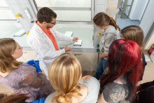 Grupa uczestników zgromadzona wokół dygestorium słucha wyjaśnień prowadzącego dotyczących wyposażenia laboratorium.