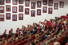 Studentki i studenci siedzą w auli i słuchają wystąpień. Na ścianie nad widownią znajdują się portrety związane z historią uczelni.