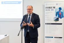 Osoba przemawia do uczestników Forum WUM-PW, stojąc przy mikrofonie. W tle widoczny jest roll-up informacyjny wydarzenia.