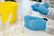 Dłonie w rękawiczkach laboratoryjnych przenoszą ciecz do probówki za pomocą pipety.