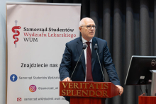 Mężczyzna przemawia przy mównicy obok roll-upu Samorządu Studentów Wydziału Lekarskiego WUM.