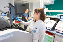 Kobieta w białym stroju medycznym i niebieskich rękawiczkach obsługuje ekran aparatury laboratoryjnej, wskazując palcem wyniki wyświetlane na monitorze. W tle stoi mężczyzna w granatowym stroju medycznym, a wokół znajdują się analizatory i sprzęt diagnostyczny.