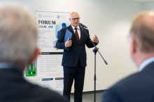 Osoba przemawia do uczestników Forum WUM-PW, stojąc przy mikrofonie. W tle widoczny jest roll-up informacyjny wydarzenia.