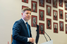 Jedna osoba przemawia z mównicy. W tle widoczna jest ściana z portretami.