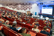 Uczestnicy konferencji siedzą w audytorium i słuchają wystąpienia prelegenta. Widoczna jest scena z mównicą oraz ekran prezentacyjny.