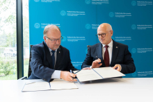 Dwie osoby podpisują dokumenty przy stole podczas spotkania w Centrum Zaawansowanych Materiałów i Technologii CEZAMAT Politechniki Warszawskiej. W tle znajduje się ścianka z logo instytucji.